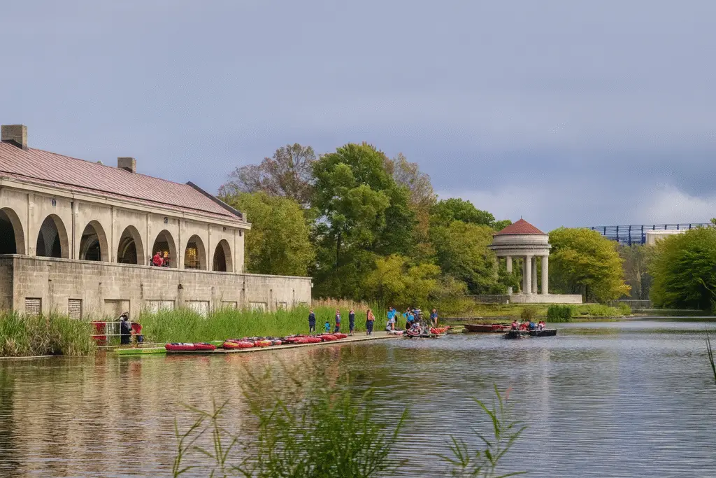 Scenic view of Fairmount Park’s boathouse along the Schuylkill River, with kayaks lined up at the dock, people enjoying water activities, and a neoclassical pavilion surrounded by lush green trees under a cloudy sky.