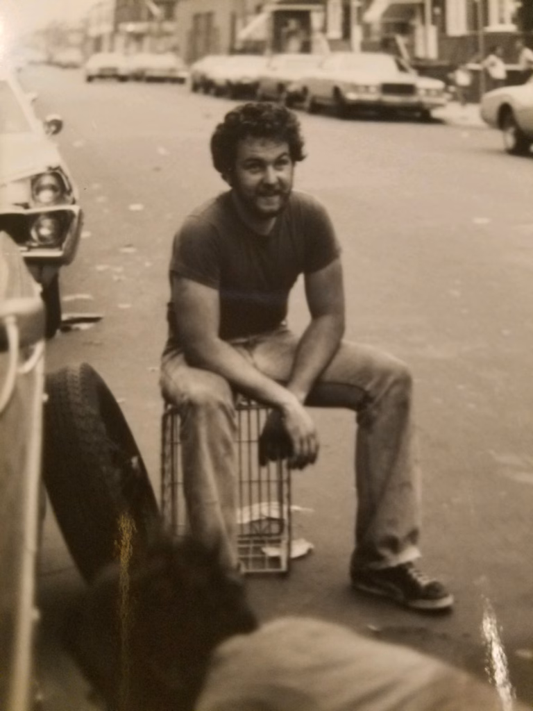 Tom Oakes sitting on a milk crate while changing a tire on Shunk Street in South Philadelphia during America's Bicentennial year in 1976.